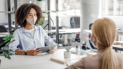 Search for workers and new vacancy after quarantine. African american protective mask woman with tablet asks blonde lady questions on interview through protective glass in modern office interior