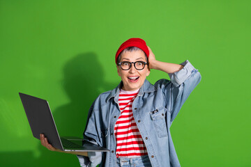 Cheerful middle aged woman in stylish denim outfit holding a laptop, expressing excitement against...