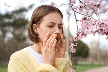 Allergic young woman sneezing near blossoming tree in park, closeup