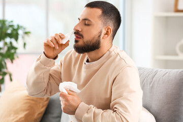 Sick young man with tissue using nasal drops from allergy at home
