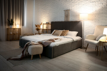 Stylish interior of bedroom with soft grey bed, comfortable armchair and glowing lamps at night