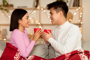 Christmas Eve. Happy Korean Couple Having Coffee Celebrating Xmas Together Sitting On Couch Covered With Blanket At Home. Winter Holidays Joy And New Year Family Leisure Concept