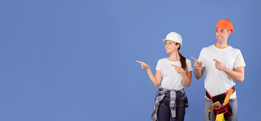 Cheerful Couple Of Workers In Hardhats And Casual Clothes Pointing Aside At Copy Space With Fingers. Positive Man And Woman In Protective Helmets Showing Free Place For Your Repair Service Ad