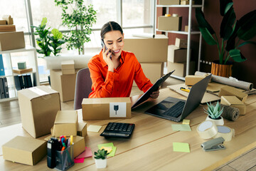 Businesswoman managing e-commerce orders at her workplace using a laptop and phone in a...