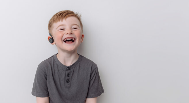 Laughing young boy with light skin, red hair, and freckles wearing hearing aid. Dressed in grey t-shirt, smiling broadly with missing teeth. Light plain background with copy space on the right

 - Powered by Adobe
