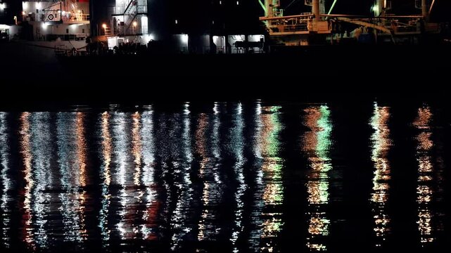 夜の波止場。船の明かりが水面に反射している
