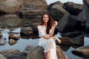 Serene woman in white dress sitting on oceanfront rock with rocks in the background