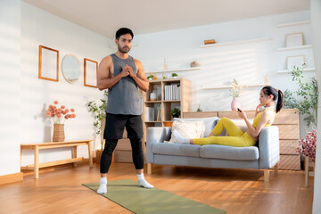 Young asian man doing squat exercise on yoga mat in living room at home while woman using phone for...