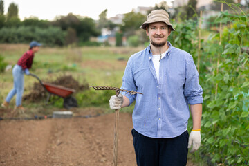 Male owner of plantation works on beds with hand tools, levels and loosens ground using rake. He...