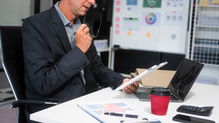 An elderly Caucasian man works at an office desk with  laptop, engaging in an online business discussion.He appears focused and professional,managing digital communication remote business operations