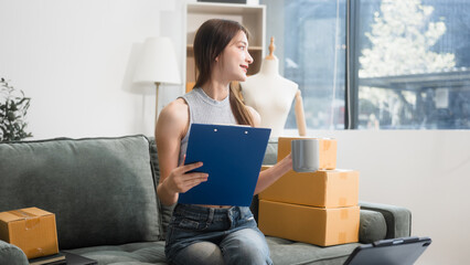 Happy Asian woman packing parcel boxes at a sofa table, smiling joyfully while checking online orders on laptop.She prepares products for delivery,managing her successful home-based online business