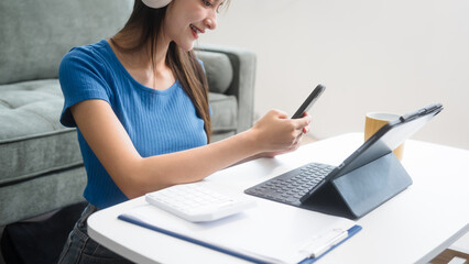 Asian woman sitting at a sofa table, wearing headphones and learning online at home.Focused on her laptop screen, she studies comfortably in a cozy environment,embracing digital education technology