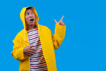 Amazed Asian man in yellow raincoat showing ads on copy space isolated on blue background © Gatot