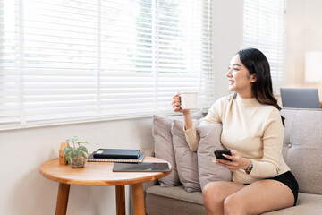 Happy young woman holding cup and mobile phone sitting on couch at home.