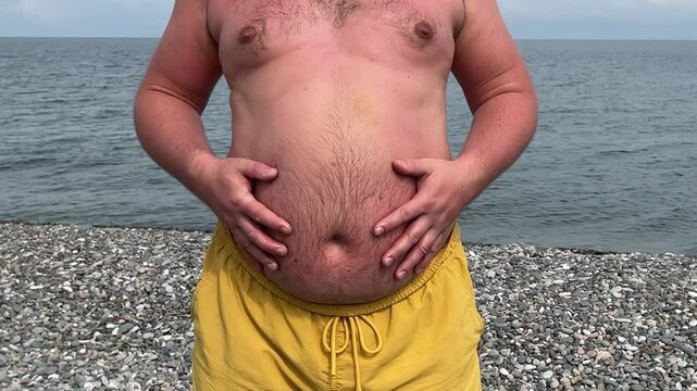 Horizontal video close up. White fat naked man in yellow shorts stands on pebble beach and slaps hands on stomach. Large hairy big belly shakes like drum. Summer vacation, tourism, walk, diet. Sun Day
