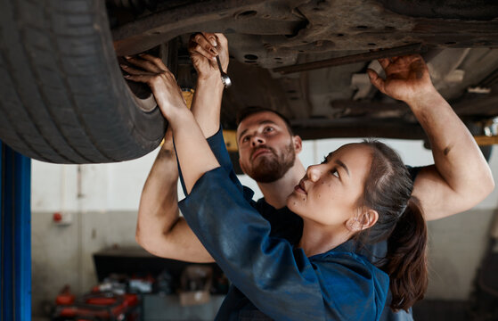 Mechanic, garage and working on car with training, maintenance engineer and teamwork at workplace. Repair shop, trade and mentor with woman checking under wheel with technician and auto service