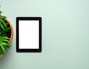 Fototapeta premium A digital tablet with a blank screen sits beside a potted green plant on a light pastel surface.