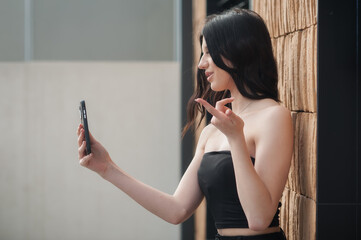 A fashionable young girl is taking a selfie in a stylish outfit against a contemporary textured wall. She strikes a playful pose, capturing the essence of social media influence during the day