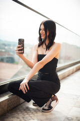 A fashionable young girl is sitting on a stylish ledge, smiling as she captures a selfie with her phone. The background shows beautiful urban scenery, indicating a relaxing afternoon