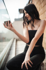 Young social media influencer captures a stylish selfie on a balcony. Dressed in a chic black outfit, she showcases her modern fashion sense while enjoying the sunny day