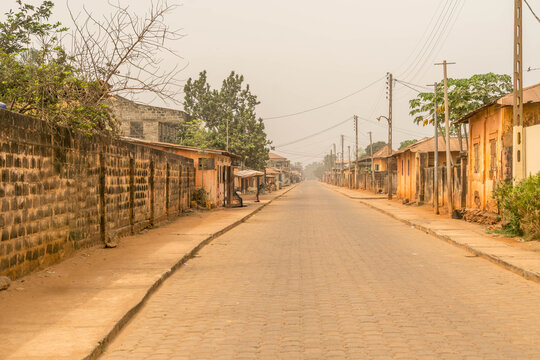 Empty cobblestone street in Ouidah, Benin, West Africa, lined with rustic buildings, trees, and power lines under a hazy sky, tranquil urban scene.
