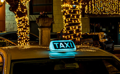  taxi with a lit-up TAXI sign on the roof. The taxi is parked in a city at holiday night, with festiveal blurry lights in the background