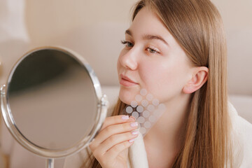 Girl examines face in mirror while holding pimple patch sheet for skincare treatment.
