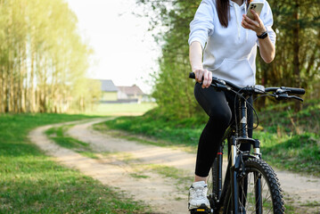Young woman riding a bicycle and holding a phone in her other hand in the forest at sunset.