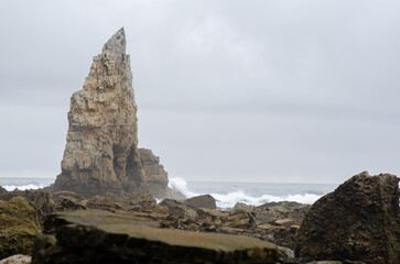 Massive sea stack rises above rocky shore with waves crashing against the cliffs on a moody,...