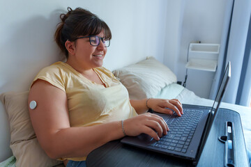 Professional woman wearing continuous glucose monitor, using laptop while resting in bed, combining...