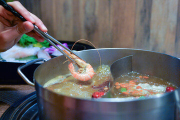 Hand using chopsticks to place a fresh shrimp into a bubbling hot pot with broth and ingredients, Asian cuisine dining concept.
