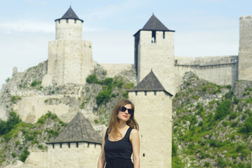 A woman with a smile on her face stands against the backdrop of stone towers and defensive walls of a medieval castle. The most impressive fortifications in Europe: Golubac Fortress in Serbia.