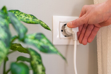 Fototapeta na wymiar Close-up of a hand inserting a white electrical plug into a white wall socket next to a green houseplant. Highlighting the concepts of electricity. Power. And energy consumption in a domestic setting
