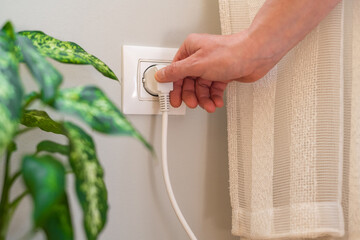 Close-up of hand inserting white electrical plug into wall socket next to houseplant, highlighting...