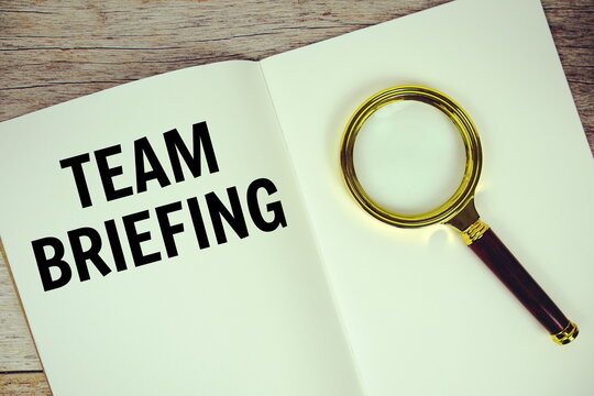 Team Briefing text message written on paper note with magnifying glass top view on wooden background - Powered by Adobe