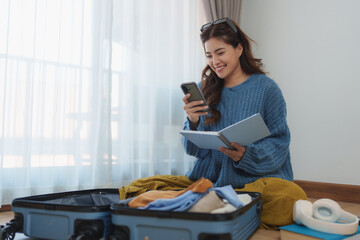 Young asian woman sitting on the floor, using her smartphone while preparing clothes for travel....