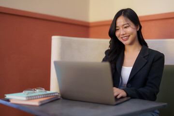 Young asian businesswoman sitting in a cafe, smiling and typing on her laptop, with notebooks and eyeglasses on the table beside her, suggesting a productive workday