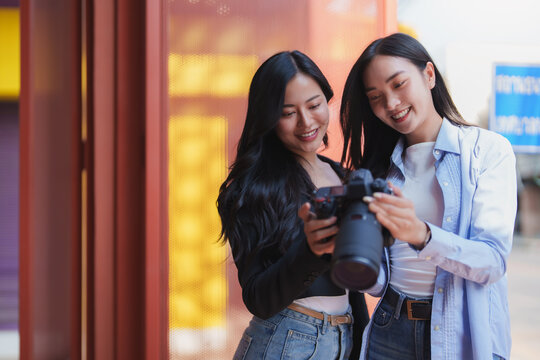 Two young female photographers are smiling while reviewing photos they've taken on their professional camera, standing in a vibrant urban environment with red and yellow background elements