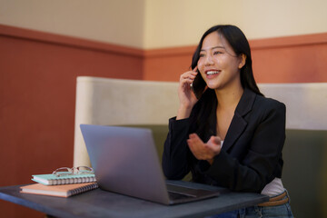 Young asian businesswoman is sitting in a cafe, talking on her phone and working on her laptop,...
