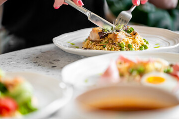 A close-up of a person cutting a piece of grilled chicken on a plate with a fork and knife. The plate contains rice, peas, and greens. Blurred background adds depth.