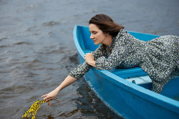 woman wearing a floral dress leans over the edge of a blue boat, delicately touching the water with...