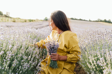 woman in yellow dress stands in lavender field, gently holding bouquet of lavender flowers. She appears serene and contemplative, enjoying beauty and fragrance of blossoms around her.