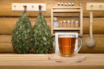 In the sauna interior, glass mug of light beer on wooden tray stands on a plank bench, against a log wall with dry birch brooms, wooden ladle and shelf with bottles of aromatic oils and white towel.