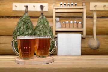 In the sauna interior, glass mugs of light beer on wooden tray stand on a plank bench, against a log wall with dry birch brooms, wooden ladle and shelf with bottles of aromatic oils and white towel.