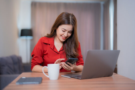 Smiling Asian businesswoman wearing red shirt using smartphone and laptop while working from home, with a cup of coffee and notebook on the table - Powered by Adobe
