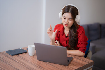 Young Asian businesswoman wearing headphones and red shirt having video call on laptop sitting at wooden table in home office, gesturing with hand explaining something