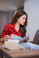 Young Asian businesswoman working from home managing domestic bills and invoices, holding documents and looking at laptop, sitting at desk with many papers and calculator