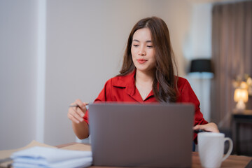 Naklejka premium Young Asian businesswoman working from home, sitting at a wooden table with a laptop computer in front of her and holding a pen, wearing a red shirt