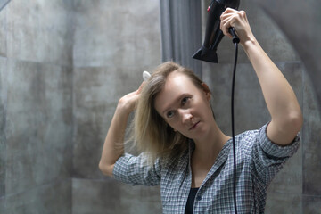 Young woman brushing her wet hair with a pink brush and drying it with a black hairdryer in a bathroom with gray tiled walls after taking a shower