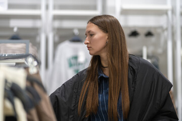 Young woman browsing through a fashion store, holding a brown pullover on a hanger and contemplating her choice for warm clothing to enhance her autumn wardrobe